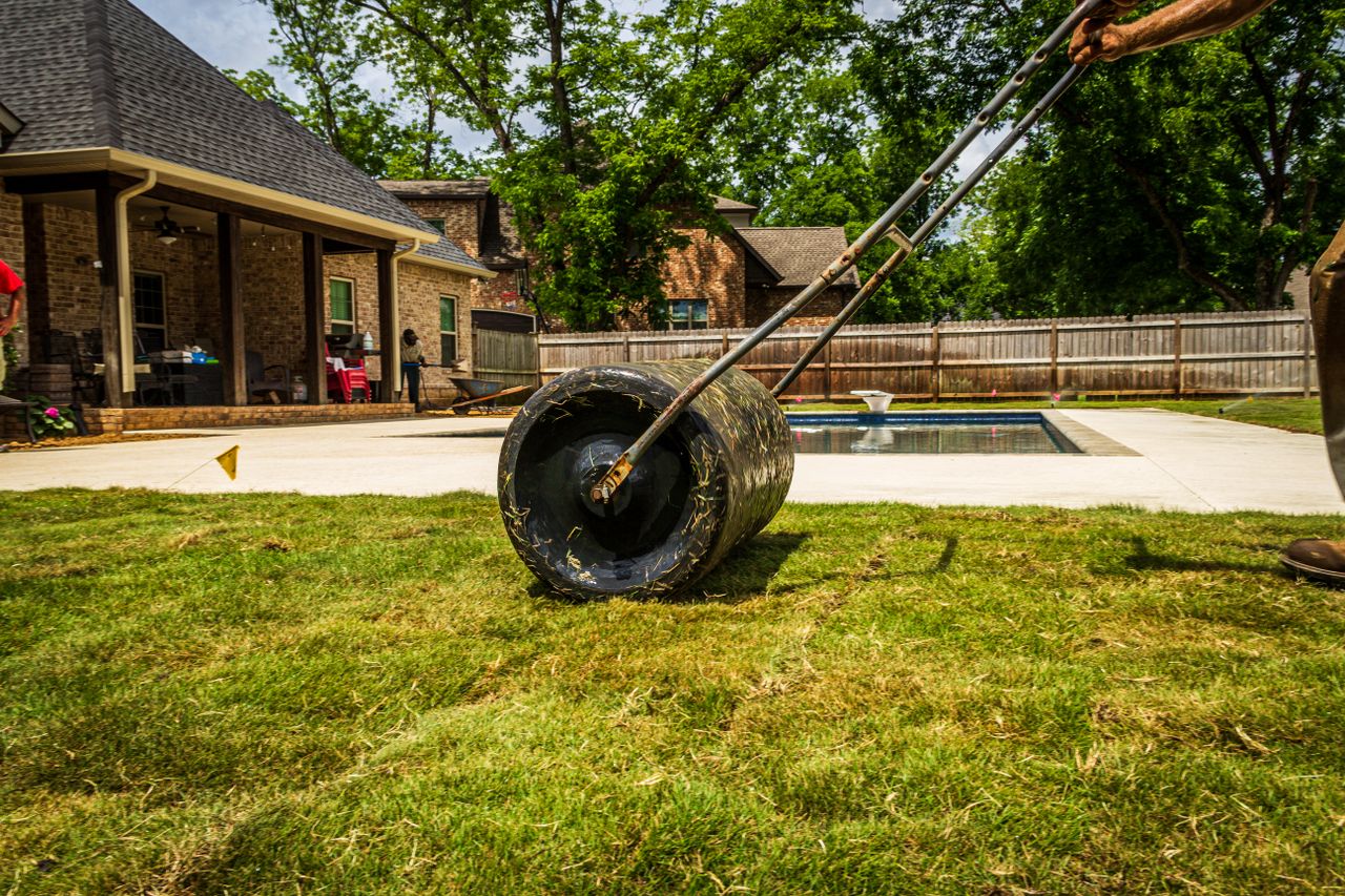 A worker rolling a lawn in a suburban backyard near a pool on a sunny day.